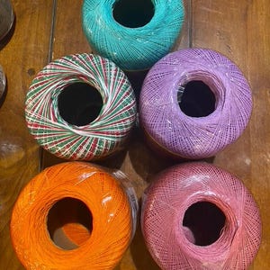 May include: Five round balls of craft thread arranged in a cluster. The colors include a bright turquoise, a solid lavender, a solid orange, a solid pink, and one ball featuring a variegated pattern of red, white, and green thin threads.