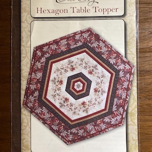 May include: A sewing pattern for a hexagon table topper titled Sew Biz Hexagon Table Topper SB 104. The cover features a hexagonal quilt design with red, cream, and dark brown floral patterns. The finished size is listed as 33 inches by 37 inches.