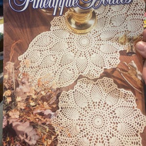 Pode incluir: Capa de livro com o título "Heirloom Pineapple Doilies" em escrita elegante. A capa apresenta toalhas de crochê creme e o texto "10 Pineapple Favorites You'll Love to Crochet!"