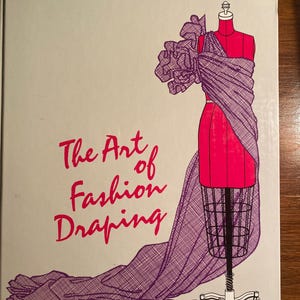 May include: Book cover with the title "The Art of Fashion Draping" in pink lettering. The cover features a purple and pink illustration of a dress form with a draped fabric garment.