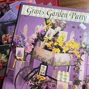 May include: A stack of books with floral and garden themes. The top book is titled "Gran's Garden Party" with a decorative cart filled with flowers. The book below is titled "Gran's Workbook". The books are by Ros Stallcup.