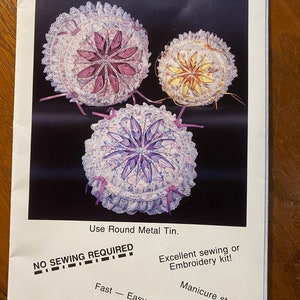 Puede incluir: Una hoja de instrucciones para un proyecto de manualidades para hacer latas decorativas con forma de margarita. La imagen muestra tres latas terminadas con flores de tela de diferentes colores. El texto en la hoja dice "Daisy Decorator Tin", "No Sewing Required", "Excellent sewing or Embroidery kit!" y "Fast - Easy - Fun."