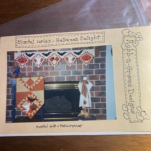 May include: A Halloween-themed quilt and table runner pattern. The image shows a mantel quilt and table runner with a brick fireplace in the background. The quilt features pumpkins, bats, and ghosts. The text reads "Mantel series - Halloween Delight."