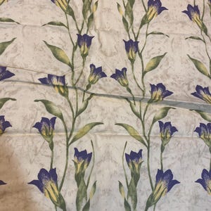 May include: Cream-colored fabric with a repeating floral pattern. The design features purple and yellow flowers with green stems and leaves. The background has a mottled, textured appearance.
