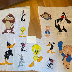 以下が含まれることがあります： Taz、シルベスター、トゥイーティーバード、ダフィーダック、バッグスバニー、ポークピッグなど、カラフルな漫画のキャラクターが描かれた白い布のパネル。キャラクターは様々なポーズで、大胆なアウトラインと鮮やかな色使いが特徴です。