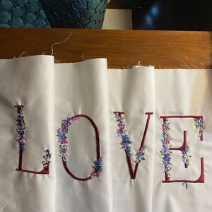 May include: Four white fabric panels with the word "LOVE" embroidered in burgundy thread. Each letter is outlined in burgundy thread and decorated with small blue and pink flowers.