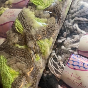 May include: Close-up of several bags of yarn in various colors. The yarn is a mix of brown, green, and white. The bags are labeled "Needle Crafters". The yarn appears to be soft and fluffy, suitable for knitting or crocheting projects.