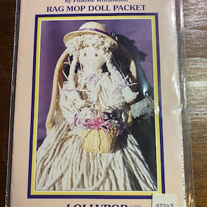 Puede incluir: Un paquete de muñeca Rag Mop, con una muñeca con sombrero de paja, sentada en una silla de mimbre, sosteniendo una cesta de flores. El paquete está etiquetado "Country Trifles" por Pauline Williamson, con "LOLLYPOP" y un aviso de copyright.