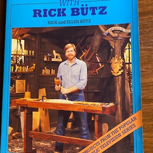 May include: A book cover with a blue background and the title "Woodcarving with Rick Butz". The cover features a photo of a man in a blue shirt carving wood at a workbench. The text "14 Projects from the Popular Public Television Series" is printed on a yellow banner at the bottom of the cover.
