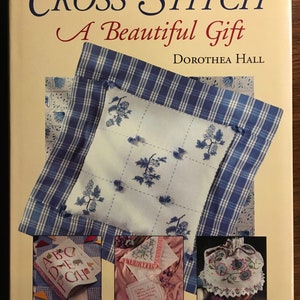 May include: Book cover with the title "Cross Stitch: A Beautiful Gift" by Dorothea Hall. The cover features a blue and white checked fabric with a white cushion embroidered with blue flowers. The book also features three smaller images of cross stitch projects: a book with the alphabet, a handkerchief with a floral design, and a drawstring bag with a floral design.