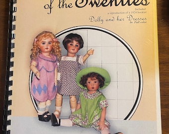 Pode incluir: Um livro intitulado "A Child of the Twenties" com uma imagem de capa de três bonecas vestindo vestidos. O livro inclui uma reprodução de um livreto de 1924 intitulado "Dolly and her Dresses" em cores. O livro é uma publicação Wish Booklet de Susan Sirkis e inclui padrões de roupas para bonecas de 28 a 30 cm de altura.
