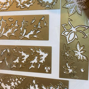 Może przedstawiać: Złote metalowe wykrojniki w r&oacute;żnych świątecznych wzorach. Obraz przedstawia kilka prostokątnych arkuszy z wyciętymi kształtami wieńc&oacute;w, liści ostrokrzewu, gwiazd betlejemskich i innych świątecznych liści. Wzory są szczeg&oacute;łowe i skomplikowane.