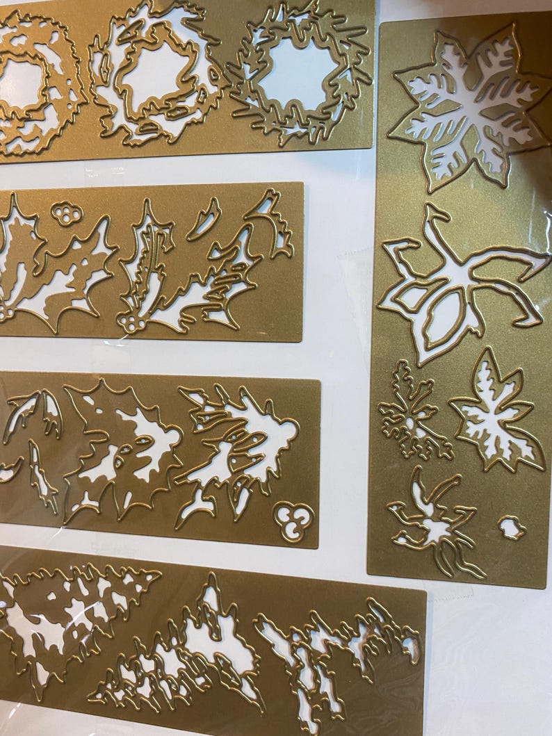 Może przedstawiać: Złote metalowe wykrojniki w r&oacute;żnych świątecznych wzorach. Obraz przedstawia kilka prostokątnych arkuszy z wyciętymi kształtami wieńc&oacute;w, liści ostrokrzewu, gwiazd betlejemskich i innych świątecznych liści. Wzory są szczeg&oacute;łowe i skomplikowane.