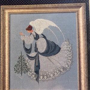 May include: Cross-stitch artwork of an angel in a gold-colored frame. The angel wears a blue and white dress with white wings and a red hairpiece. A small green tree is on the left. The background is a light blue color.