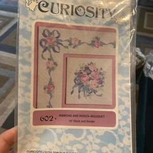 May include: A vintage cross-stitch pattern featuring a bouquet of pink and purple roses with blue ribbons. The pattern is titled "Ribbons and Roses-Bouquet" and is 16 inches in size. The pattern is from Curiosity, a company located in Monument, Colorado.