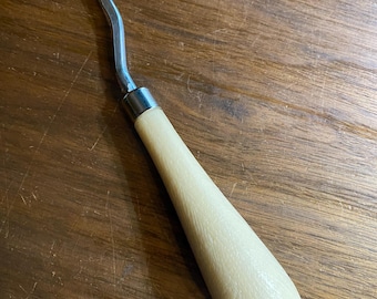 May include: A metal latch hook tool with a curved, hooked end and a light-colored wooden handle. The tool is designed for rug hooking, latch hooking, and other craft projects. The handle is smooth and tapered.