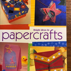 May include: A book titled "Bright ideas in papercrafts" with a colourful cover featuring a variety of papercraft projects, including a box decorated with buttons and yarn, a star ornament, a baby bib card, and a red envelope with a tassel. The book includes 23 projects.