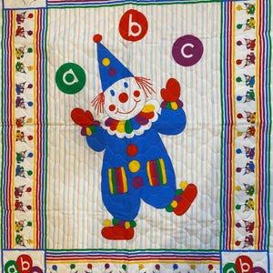 May include: A vibrant, quilted baby blanket with a playful clown motif. The clown is in blue with red details and red gloves. The blanket has a striped border with balloons and the letters a, b, and c. The background is white with vertical stripes.