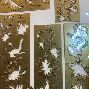 Może przedstawiać: Kolekcja złotych wykrojnik&oacute;w w kształcie kwiat&oacute;w i liści. Kształty ułożone są na prostokątnym i kwadratowym złotym kartonie. Wycięte wzory odsłaniają białą przestrzeń, tworząc kontrast między złotymi i białymi elementami. Obraz przedstawia r&oacute;żne wzory botaniczne.