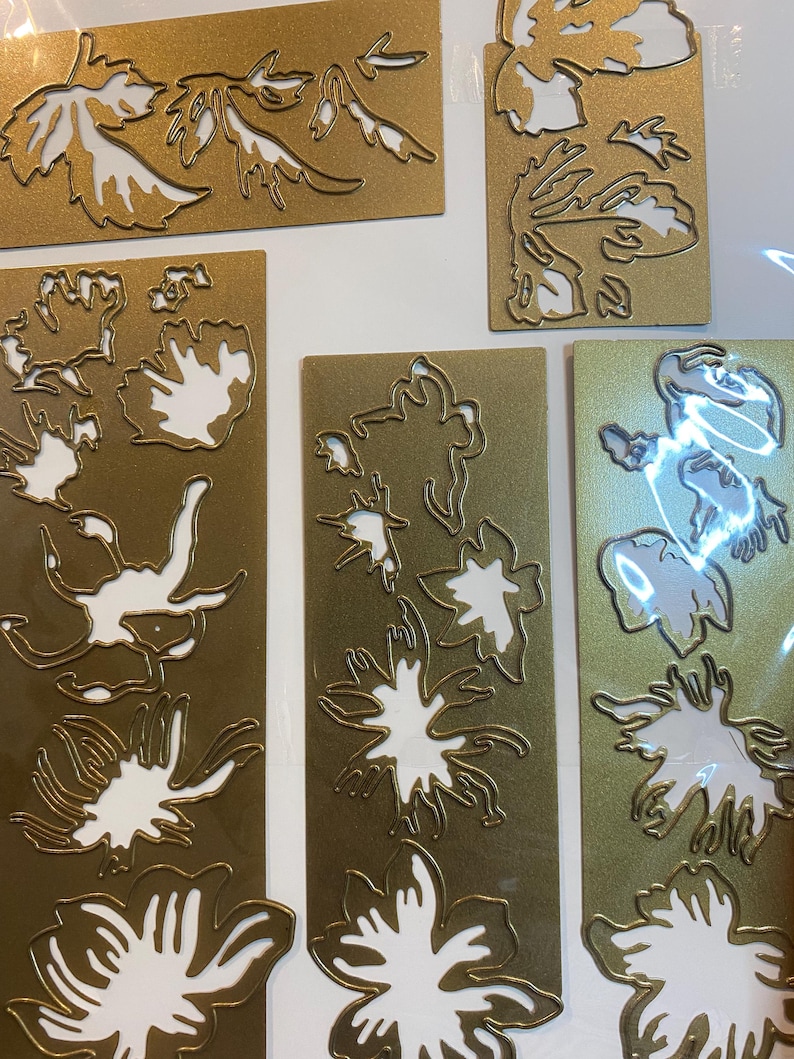 Może przedstawiać: Kolekcja złotych wykrojnik&oacute;w w kształcie kwiat&oacute;w i liści. Kształty ułożone są na prostokątnym i kwadratowym złotym kartonie. Wycięte wzory odsłaniają białą przestrzeń, tworząc kontrast między złotymi i białymi elementami. Obraz przedstawia r&oacute;żne wzory botaniczne.