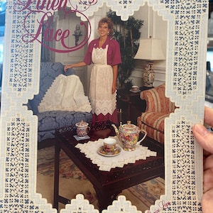 May include: A magazine cover titled "Linen & Lace Hardanger" featuring a woman in a red top and white apron. The cover has a blue border with a lace design. A table is set with a teapot, teacup, and doily.