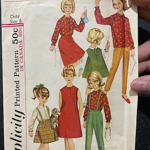 Könnte beinhalten: Simplicity Schnittmuster Nummer 5604 für Kinderkleidung. Das Schnittmuster enthält fünf verschiedene Designs für Mädchenkleider, Röcke, Hosen und Oberteile. Das Schnittmuster ist auf weißem Hintergrund mit roten, grünen und braunen Illustrationen gedruckt.