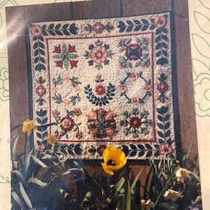 Puede incluir: Un tapiz de pared acolchado con diseños florales en rojo, rosa y verde sobre un fondo crema. El edredón presenta un borde de hojas verdes oscuras y se exhibe contra un fondo de madera, con tulipanes amarillos en primer plano.