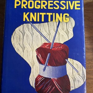 May include: A hardcover book titled The Complete Book of Progressive Knitting by Ida Riley Duncan. The blue cover features a graphic illustration of a red yarn ball with knitting needles, set against a cream-colored textured cable knit pattern.