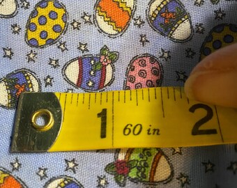 Pode incluir: Tecido azul claro com um padrão repetido de ovos de Páscoa em várias cores e designs, incluindo bolinhas amarelas, listras laranja e detalhes florais. Uma fita métrica amarela mostra 152 cm.