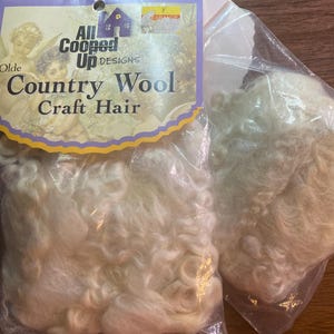 May include: Two clear plastic bags containing off-white, curly wool craft hair. The packaging features a label with the text All Cooped Up Designs Olde Country Wool Craft Hair and an illustration of two cherubs. A small yellow clearance sticker is visible.