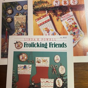 May include: A collection of Christmas-themed cross-stitch pattern books. The covers feature images of Christmas stockings, ornaments, and festive designs. Titles include "Christmas," "Yuletide Bears," and "Frolicking Friends." The books showcase holiday-themed cross-stitch projects.