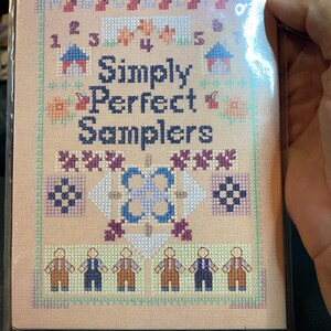 May include: A pink and white cross-stitch sampler with the title "Simply Perfect Samplers". The sampler features a variety of designs, including letters, numbers, houses, flowers, and a central design of a blue and white flower.