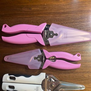 May include: Three pairs of gardening shears are displayed. Two pairs are pink with clear plastic blade covers. The third pair has white and black handles with a silver blade. The shears are designed for trimming plants and flowers.
