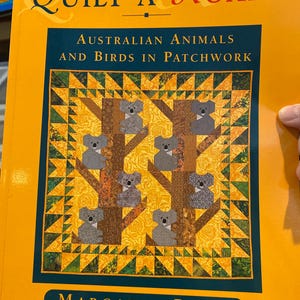 以下が含まれることがあります： 「Quilt a Koala」というタイトルの本で、黄色い表紙です。表紙には、木にいるコアラのパッチワークキルトのデザインが施されています。本の副題は「オーストラリアの動物と鳥のパッチワーク」で、著者はマーガレット・ロルフです。