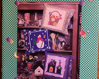 Könnte beinhalten: Ein Buch mit dem Titel "Calendar Patches for Pillows, Quilts, and Clothing" von Nancy J. Smith & Lynda S. Milligan. Der Umschlag zeigt einen grün-weiß karierten Hintergrund mit einem Bücherregal, in dem drei Kissen mit verschiedenen Designs ausgestellt sind. Die Kissen sind mit einem Blumenkorb, einem Schneemann und einem Vogelhaus verziert.