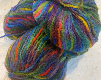 May include: Three balls of vibrant yarn in shades of purple, green, red, and blue. The yarn has a textured appearance, with visible strands and a slightly fuzzy texture. The balls are stacked, with some loose yarn strands visible.
