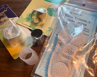 May include: Assortment of thimbles, including a silver ring thimble, a clear plastic thimble, a metal thimble, and a leather thimble. The leather thimble is labeled "Skin Thimble" and "Model ST3538".