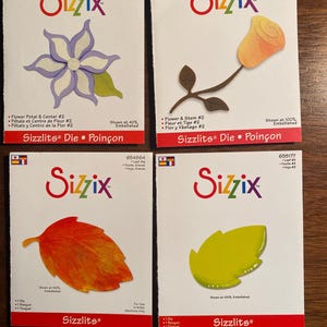 以下が含まれることがあります： 白いパッケージに収められた4つのSizzixダイカットセット。花柄と葉柄のデザインが特徴です。セットには、紫と白の花、オレンジ色のバラ、オレンジ色の葉、緑色の葉が含まれています。各パッケージには、Sizzixのロゴと製品の詳細が表示されています。