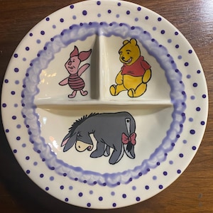 Può includere: Un piatto bianco diviso con illustrazioni dei personaggi di Winnie the Pooh. Il piatto ha un bordo a pois blu e un anello dipinto di blu. Pimpi, Winnie the Pooh ed Eeyore sono raffigurati in sezioni separate.