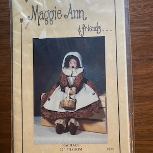 May include: A sewing pattern for a 22 inch doll named Rachael Pilgrim. The pattern is titled "Maggie Ann & Friends..." and features a brown-haired doll wearing a brown dress with a white apron and a basket. The pattern number is #260.