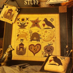 Puede incluir: Un patrón de punto de cruz con una variedad de imágenes de temática rural, que incluyen una colmena, un corazón, un cuervo y una luna. El patrón está enmarcado en un marco de madera y se muestra sobre un mantel a cuadros. El título del patrón es "Neat Country Stuff" y el diseñador es Sandra Sullivan.