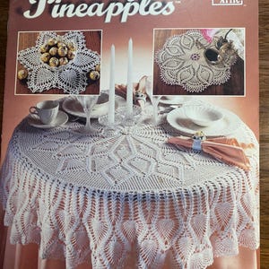Puede incluir: Libro de patrones de crochet titulado "Pretty Pineapples" de Annie's Attic. La portada del libro presenta un mantel de crochet blanco con un diseño de piña, dos manteles individuales de crochet blanco y un tazón de caramelos de chocolate.