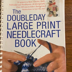 Puede incluir: El libro de labores de aguja Doubleday en letra grande con más de 100 ilustraciones fáciles de seguir. La portada del libro presenta un primer plano de una mano tejiendo con agujas e hilo.