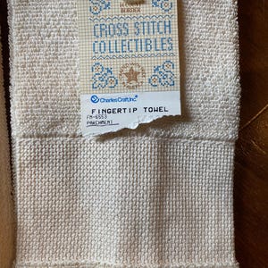 Puede incluir: Primer plano de una toalla de mano crema con un tejido texturizado. Una etiqueta dice "CROSS STITCH COLLECTIBLES" con dise&ntilde;os de punto de cruz azules y "FINGERTIP TOWEL". La toalla est&aacute; etiquetada como "PARCHMENT".