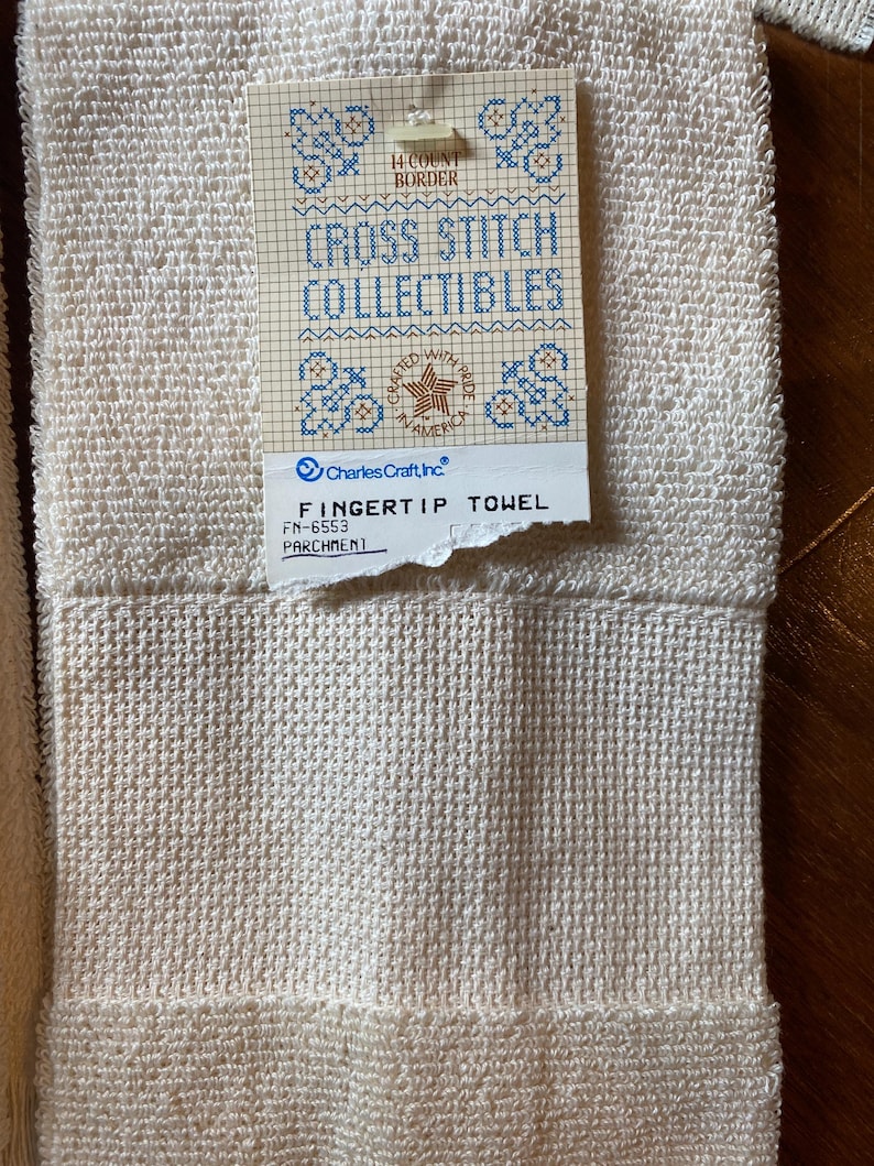 Puede incluir: Primer plano de una toalla de mano crema con un tejido texturizado. Una etiqueta dice "CROSS STITCH COLLECTIBLES" con dise&ntilde;os de punto de cruz azules y "FINGERTIP TOWEL". La toalla est&aacute; etiquetada como "PARCHMENT".