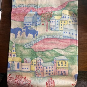 May include: A fabric runner featuring a repeating pastoral scene with blue cows and horses, yellow and pink houses, green rolling hills, and red trees. The design includes stone walls and a church steeple, rendered in a sketch-like illustration style.