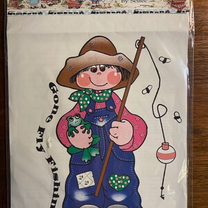 May include: Iron-on transfer featuring a cartoon girl with brown hair wearing a brown hat, pink shirt, blue overalls, and holding a fishing pole. She has a green frog on her shoulder. The text "Gone Fishin'" is written in a curved font on the left side of the image.
