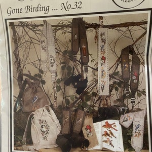 May include: A cross stitch pattern for a camera strap featuring a bird and floral design. The pattern is titled "Gone Birding... No. 32" and is designed by Paula Minkebige. The pattern is from the Crossed Wing Collection.