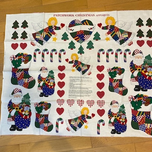 以下が含まれることがあります： クリスマスをテーマにしたパッチワークアップリケが施された白い生地。天使、サンタクロース、クリスマスストッキング、キャンディケイン、ハート、クリスマスツリーのデザイン。上部には「PATCHWORK CHRISTMAS APPLIES」の文字が印刷されています。
