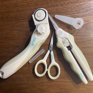 Può includere: Un set di strumenti per l'artigianato color crema e bianco, tra cui un taglierino rotante, forbici e un rifinitore, disposti su una superficie di legno. Il marchio "We R" è visibile sugli strumenti.